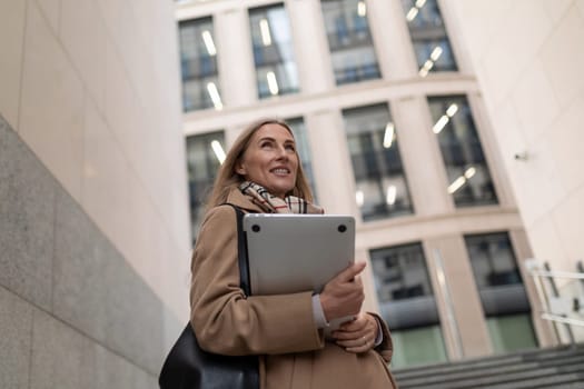 A professional woman is walking near a sleek business center. She carries a laptop and smiles, embodying confidence in a bustling urban environment.