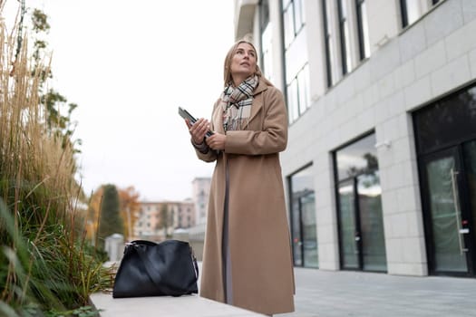 A businesswoman in a tan coat stands outside a business center. She is holding a tablet in her hand and looks up at the building, seemingly contemplating her next step.