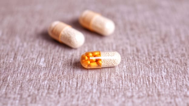 Tiny yellow medicines are spilling out of an open capsule laying on a table with two other capsules in the background out of focus