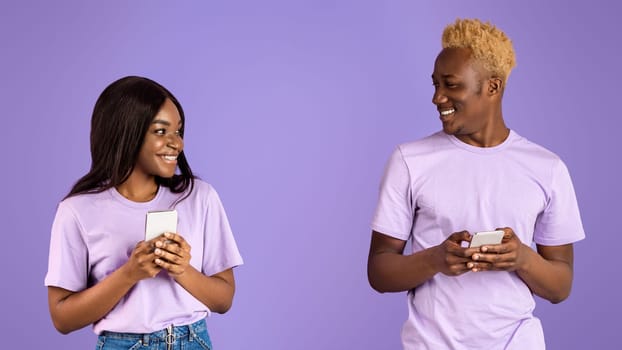 Modern communication concept. Portrait of millennial black couple with cellphones looking at each other over lilac background