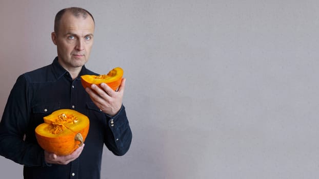 A handsome elderly man in a blue shirt against the wall holds a cut orange pumpkin with his hands and looks at the camera.