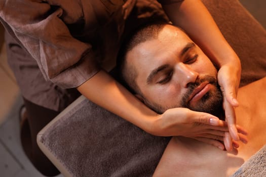 Masseuse giving man facial massage in calm relaxing atmosphere, relaxed man lying on massage table with closed eyes in spa salon