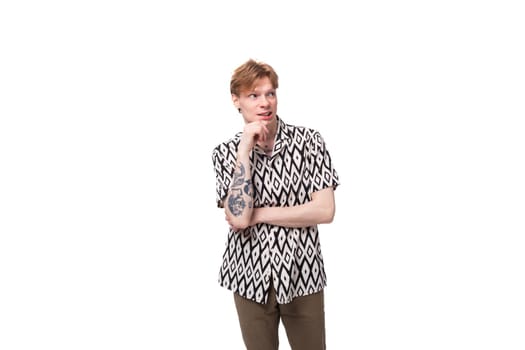 portrait of a pensive smart young informal guy with red hair and a tattoo on his arms is dressed in a short-sleeved shirt with a pattern.