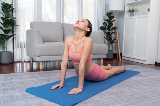 Asian woman in sportswear doing yoga exercise on fitness mat as her home workout training routine. Healthy body care and calm meditation in yoga lifestyle with comfortable and relaxation. Vigorous