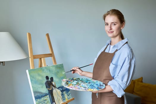 Young pretty female artist painting with acrylics on easel. Teenage girl is student of art school courses, drawing at home. Creativity hobby education, art therapy, paintings for sale, youth concept
