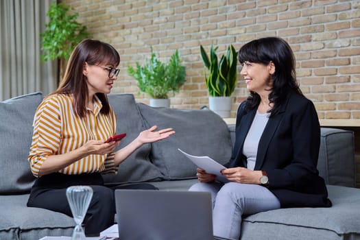 Two mature business women having conversation sitting on couch in office, discussing work, commercial projects. Communicating female colleague, 40s 50s age. Law finance mentoring consulting teamwork