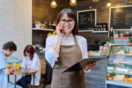 Portrait of confident middle aged woman coffee shop worker. Female in apron holding clipboard with orders looking at camera talking on cell phone with cafeteria customers. Small business work people