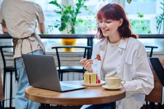 Female college student with laptop in cafeteria at table with cup of coffee and piece of cake. Internet online technology for leisure communication blogging learning chat, youth lifestyle concept