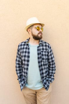 Handsome millennial man wearing summer hat and plaid shirt smiling happy near wall - travel vacations and summer holiday