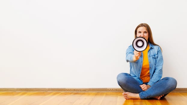 Young beautiful woman screaming on a megaphone sitting on a wooden floor in front of white wall.