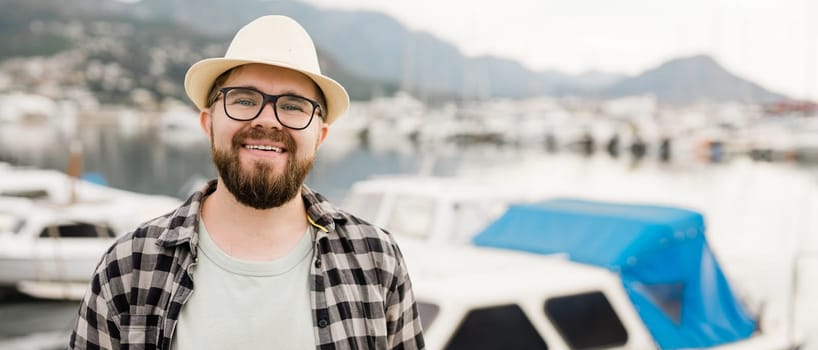 Banner millennial man wearing hat and glasses near marina with yachts. Portrait of laughing man with sea port background with copy space.