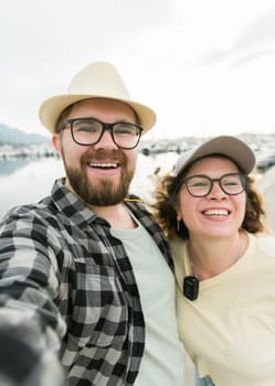 Young couple taking a self portrait laughing as they pose close together for camera on their smartphone outdoors in summer port with boats and yachts.
