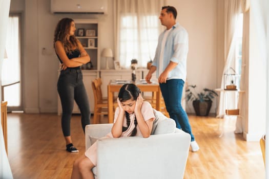 Annoyed and unhappy young girl sitting on sofa trapped in middle of tension by her parent argument in living room. Unhealthy domestic lifestyle and traumatic childhood develop to depression.Synchronos