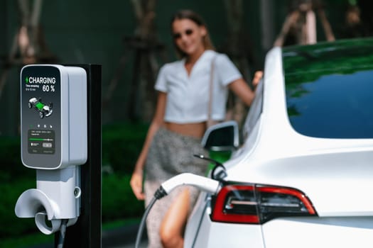 Young woman travel with EV electric car charging in green sustainable city outdoor garden in summer. Urban sustainability lifestyle by green clean rechargeable energy of electric BEV vehicle innards