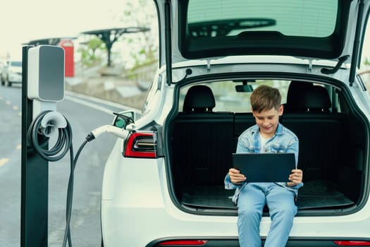 Little boy sitting on car trunk, using tablet while recharging eco-friendly car from EV charging station. EV car road trip travel as alternative vehicle using sustainable energy concept. Perpetual