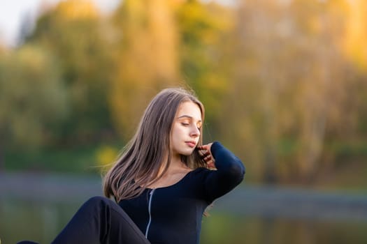 A beautiful girl poses while sitting on the grass by a pond in an autumn park