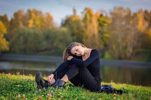 A beautiful girl poses while sitting on the grass by a pond in an autumn park