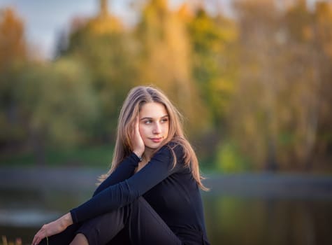 A beautiful girl poses while sitting on the grass by a pond in an autumn park