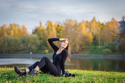 A beautiful girl poses while sitting on the grass by a pond in an autumn park