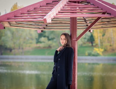 A beautiful girl poses while standing by a pond under an umbrella in an autumn park