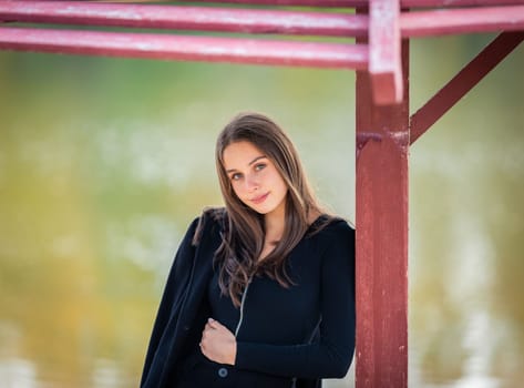 A beautiful girl poses while standing by a pond under an umbrella in an autumn park