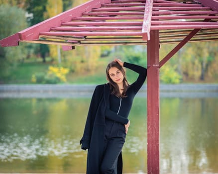 A beautiful girl poses while standing by a pond under an umbrella in an autumn park