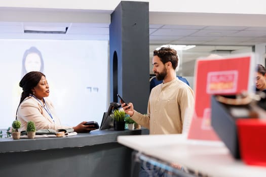 Male customer holding smartphone making contactless payment while shopping for clothes in fashion boutique. Young man standing at clothing store cashier counter using mobile phone to pay for purchases