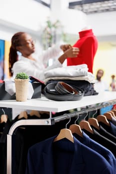 Jackets hanging on rack and accessories on shelf in clothing store while assistant dressing mannequin on blurred background. Male blazers and belts in department shopping mall