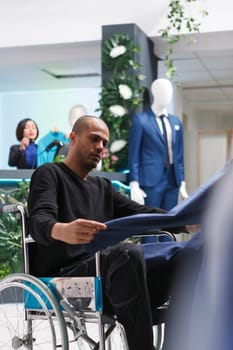 Clothing store arab client with physical impairment checking jacket style while selecting formal apparel. Inclusive boutique young customer in wheelchair exploring garment rack