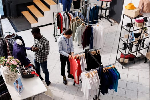 Asian employee preparing boutique for opening, arranging trendy merchandise on hangers in shopping centre. Man wearing formal shirt looking at racks full with new fashion collection in clothing store