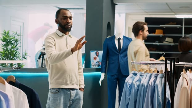 African american man looking at hologram in store, using augmented reality holographic image in fashion clothing boutique. Male customer checking clothes in shopping center. Tripod shot.