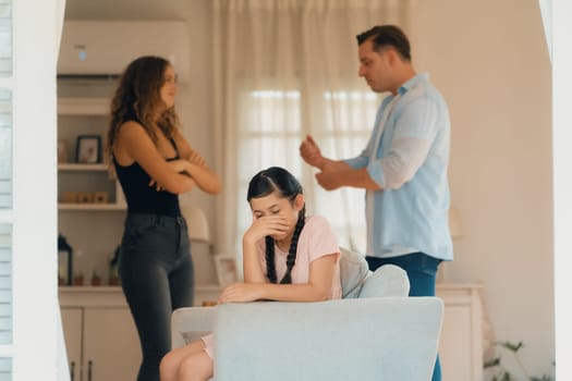 Annoyed and unhappy young girl sitting on sofa trapped in middle of tension by her parent argument in living room. Unhealthy domestic lifestyle and traumatic childhood develop to depression.Synchronos