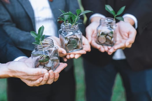 Concept of sustainable money growth investment with glass jar filled with money savings coins with businesspeople as eco-friendly financial investment nurtured with nature and healthy retirement. Gyre