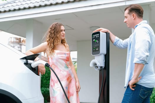 Happy and lovely couple with eco-friendly conscious recharging electric vehicle from EV home charging station. EV car technology utilized for residential home to future sustainability. Synchronos