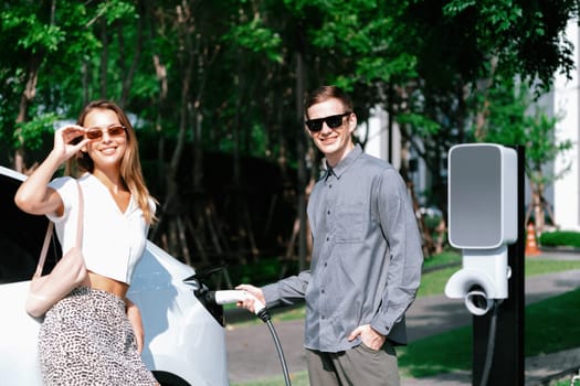 Young couple travel with EV electric car charging in green sustainable city outdoor garden in summer shows urban sustainability lifestyle by green clean rechargeable energy of electric vehicle innards