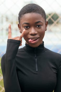 smiling young african american female showing tongue and demonstrating horns gesture with hand in town on summer day