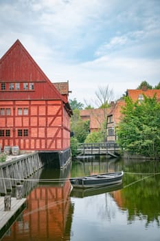 Charming red timber house by a peaceful canal, reflecting traditional Danish architecture. Surrounded by lush greenery and tranquil waters, this serene scene with watermill in the background