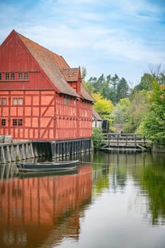Charming red timber house by a peaceful canal, reflecting traditional Danish architecture. Surrounded by lush greenery and tranquil waters, this serene scene with watermill in the background