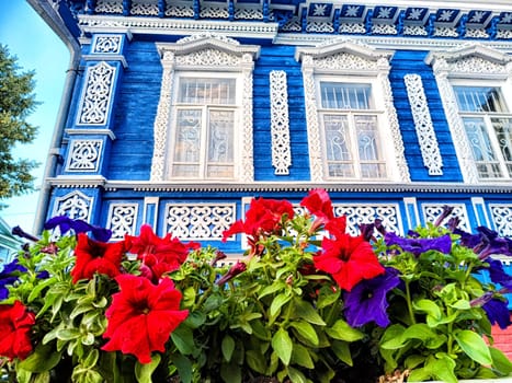 Colorful flowers in red and purple thrive in a garden while a stunning blue wooden home with ornate carvings stands majestically nearby.