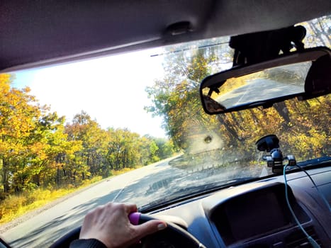 A person grips the steering wheel while driving down peaceful highway lined with autumn foliage and bright sunshine
