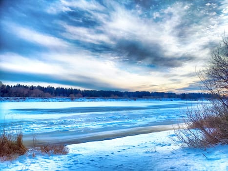 The river is partially frozen, reflecting the vibrant sunset while snow blankets the banks and trees stand in silhouette.