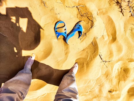 A vibrant blue sandal lies in warm sand while a person's shadow and bare feet are visible in a sunlit desert landscape.