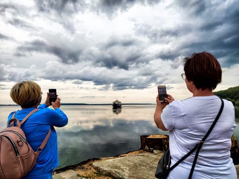 Standing by the lake, woman uses her phone to photograph the serene water and a boat against a backdrop of clouds