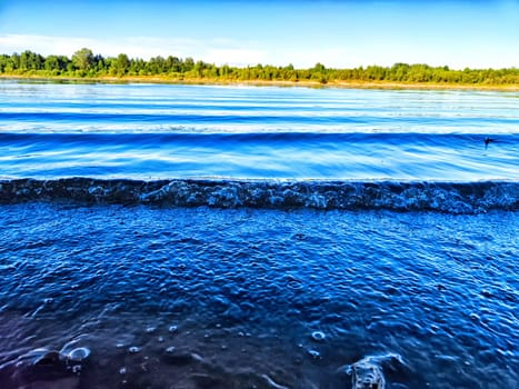 Gentle waves roll onto the rocky shore, reflecting the open blue sky and lush greenery nearby.