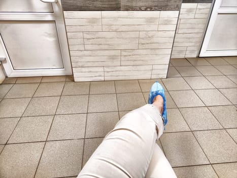 A person is waiting in a doctor's clinic hallway, sitting with feet in blue shoe covers on tiled flooring