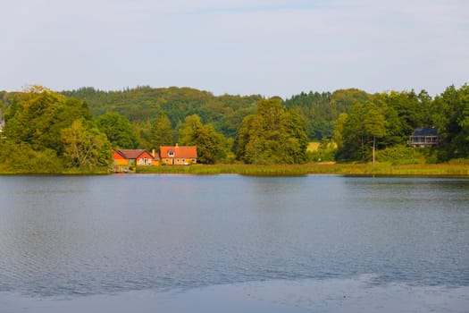 Serene landscape featuring calm waters, lush greenery, and picturesque orange wooden houses by the lakeside, evoking tranquility and natural beauty. Ideal for portraying rural life and peaceful retreats in denmark