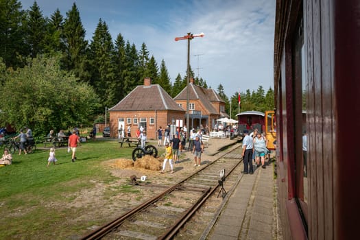 vrads,denmark,08-09-20-24:People enjoy a day at the steam train station vrads,surrounded by nature, with families and friends exploring the picturesque setting. Trains, trees, and a brick building create a nostalgic atmosphere