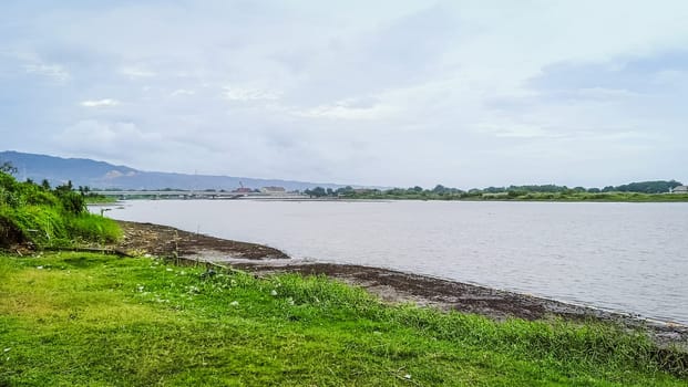 A river with a bridge in the background. The water is calm and the grass is green. The scene is peaceful and serene