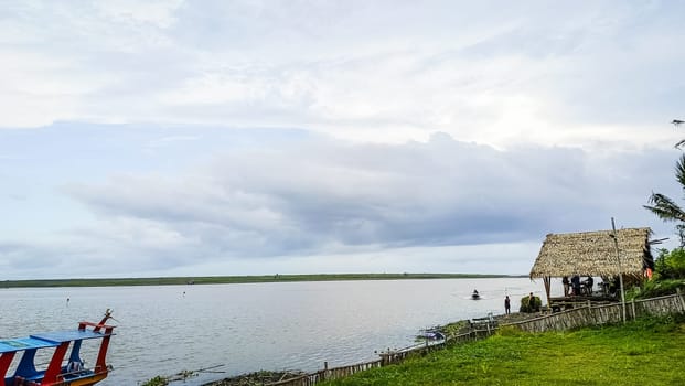 A beach with a small hut and a boat in the water. The sky is cloudy and the water is calm
