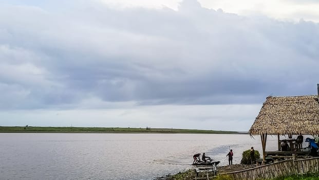 A beach with a small hut and a boat in the water. The sky is cloudy and the water is calm